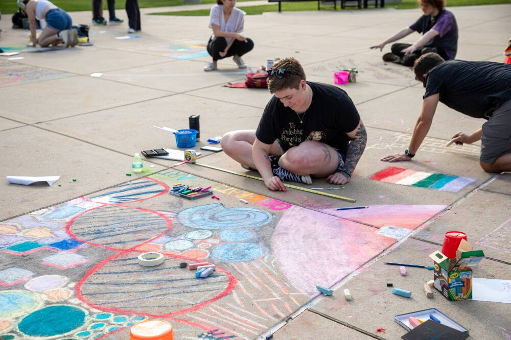 A person sits cross-legged on a concrete pavement using colored chalks to draw a large, geometric sidewalk painting in pastel shades, while other artists kneel in the background creating similar chalk artworks.